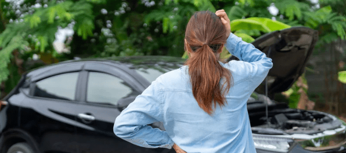 A worried woman holds her head while staring at her damaged car.